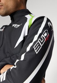 Black athletic jacket with white and green accents, featuring a high collar, zipper closure, and branding on the sleeve. Smooth texture.