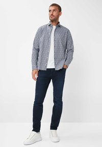 Musterhemd mit langen Ärmeln in Blau und Weiß mit Kentkragen, getragen über einem weißen T-Shirt. Kombiniert mit dunkelblauen Jeans und weißen Sneakers.