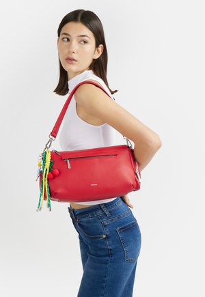 Giovane donna con top bianco senza maniche e jeans blu porta una borsa a tracolla rossa con accessori con nappe colorate.