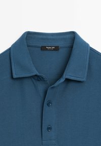 Modrá polo košile s texturovanou tkaninou, s límečkem a tříknoflíkovým zapínáním. Štítek říká "Massimo Dutti." Vyrobeno v Číně.