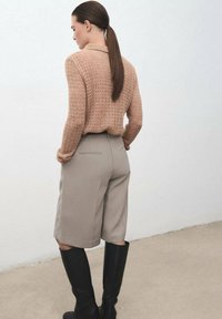 Suéter de punto marrón claro con un patrón texturizado, combinado con pantalones cortos beige a medida. Las botas negras hasta la rodilla completan el atuendo.