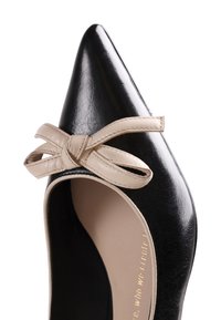 Chaussure plate noire à bout pointu avec un détail de nœud en cuir beige sur le dessus, montrant partiellement l'intérieur et le texte de la marque à l'intérieur.