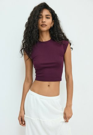 Ung kvinde med langt krøllet hår iført en ærmeløs lilla croptop og et hvidt skørt, der står foran en ensfarvet hvid baggrund.
