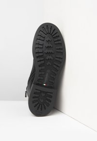 Suela de bota negra con patrón de relieve profundo, pequeño detalle de la bandera italiana y marcado de talla 42, posicionada en vertical contra muros blancos.