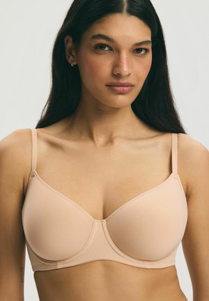 Woman with long dark hair wears a beige underwire bra, facing forward with neutral expression against a plain light background.
