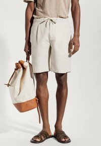 Shorts en lin beige avec une taille élastique et un cordon de serrage ; accompagnés d'un sac en toile avec accents en cuir marron clair et de sandales marron à enfiler.