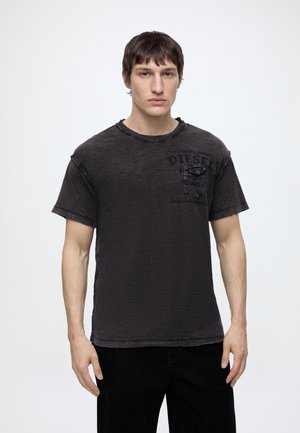 Mężczyzna z krótkimi brązowymi włosami ma na sobie wyblakły czarny t-shirt Diesel z małą kieszonką na piersi, stojąc na tle jednolitego białego tła.