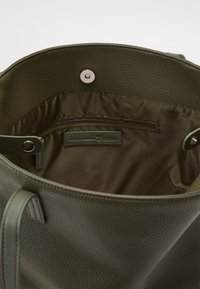 Sac vert olive avec un extérieur texturé et granuleux. L'intérieur présente une doublure en tissu, une poche zippée et une étiquette de marque sur une surface lisse.