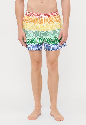 Mann trägt buntes Badebekleid mit dem sich wiederholenden Schriftzug "SQUARED2" in Regenbogenstreifen und steht barfuß vor weißem Hintergrund.