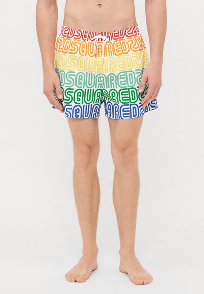 Homme portant un short de bain multicolore avec le texte "SQUARED2" répété en bandes arc-en-ciel, se tenant pieds nus sur fond blanc.