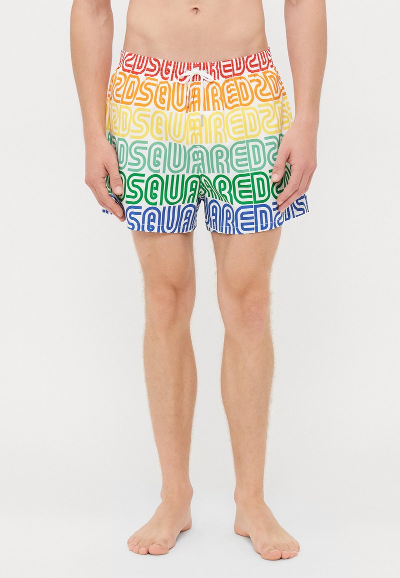 Man som bär flerfärgade badshorts med upprepande "SQUARED2"-text i regnbågsränder, stående barfota mot en vit bakgrund.