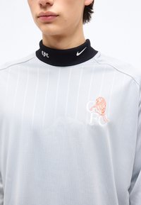 Νεαρός που φοράει ανοιχτογρι ζακέτα με κάθετες ρίγες και μαύρο ψηλό γιακά με λογότυπα "CFC" και Nike.