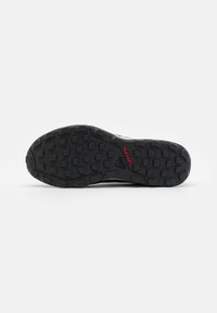 Suola di scarpa in gomma nera con un motivo di presa testurizzato, caratterizzata da lobbete rettangolari rialzate. "TRAXION" e logo del marchio in rosso vicino al tallone.