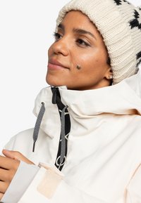 Helle wetterfeste Jacke mit hohem Kragen, schwarzen Kordeln und einem Klettverschlussbund. Strukturierte Strickmütze mit schwarzen Akzenten.