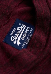 Tissu texturé rouge foncé avec une étiquette de marque SuperDry en bleu marine cousue à l'intérieur, présentant un texte blanc et des caractères japonais.