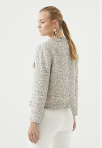 Grijze en witte getextureerde tweedjas met franjes, getailleerde pasvorm en lange mouwen, gedragen over witte broeken.