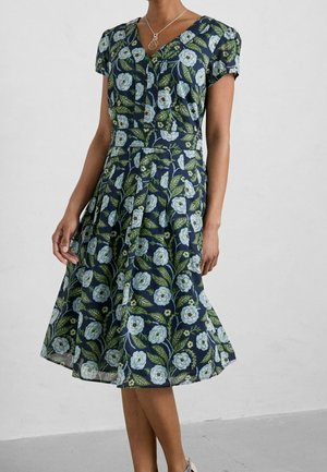 Femme portant une robe bleu marine mi-longue à imprimé floral bleu clair, manches courtes, col en V, et un collier argenté sur un fond uni.