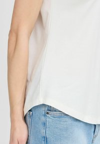 Camicia di cotone bianco con orlo curvo, abbinata a jeans in denim blu. I tessuti sembrano morbidi, con cuciture visibili e una vestibilità rilassata.