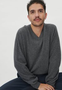 Grauer V-Ausschnitt-Pullover aus weichem Strickstoff mit langen Ärmeln, der auf einer dunklen Hose liegt. Auffällig durch seine lockere Passform und leichte Textur.