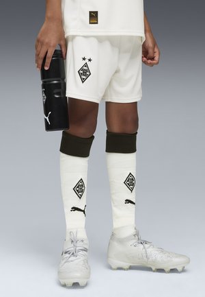 Piernas en uniforme de fútbol blanco con logotipos negros, sosteniendo una botella de agua negra de Puma, usando zapatillas blancas y calcetines blancos con logotipos negros.