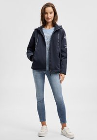 Marineblaue Jacke mit Kapuze, Reißverschluss und verstellbaren Kordeln; getragen über einem hellblauen T-Shirt mit Grafik. Kombiniert mit hellblauen Jeans und weißen Turnschuhen.