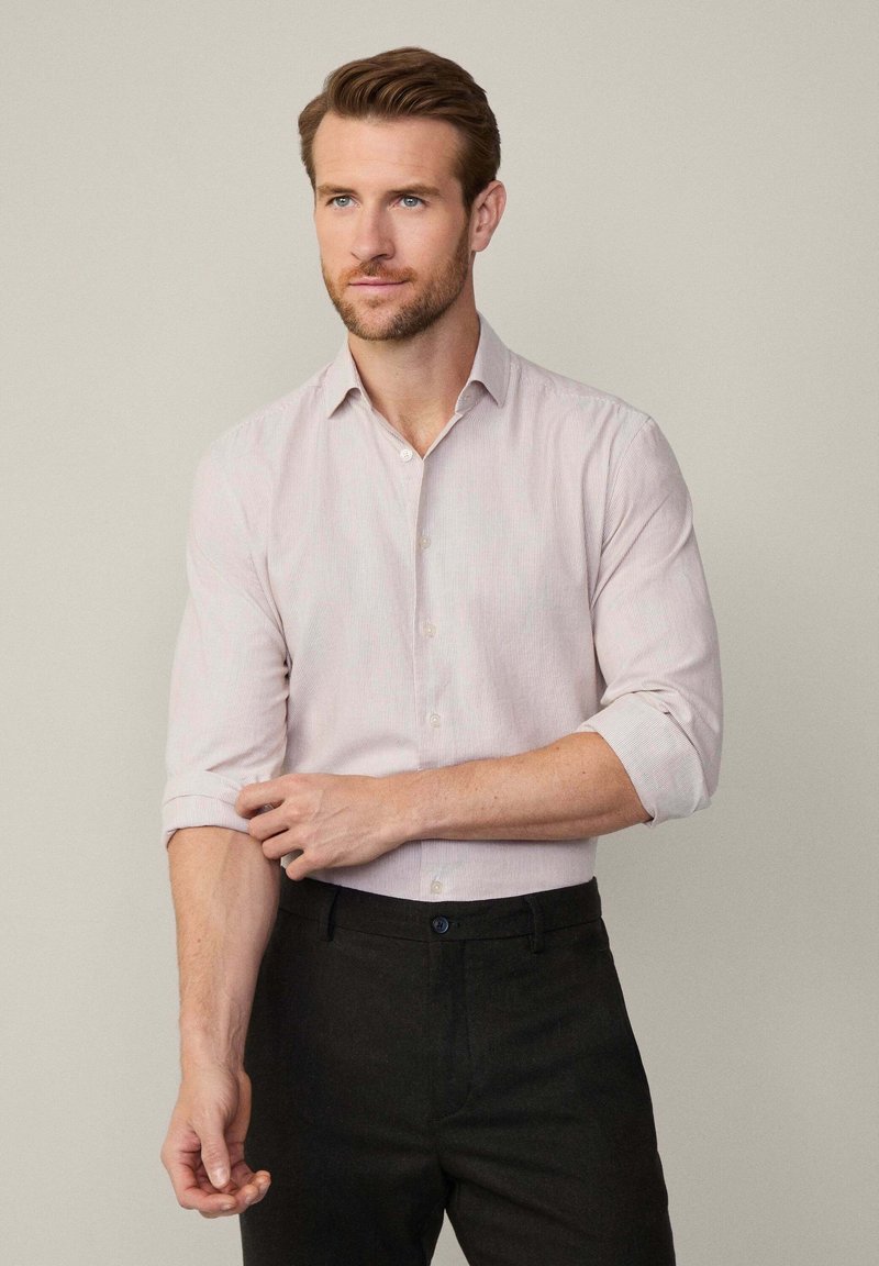 Camisa de botón a rayas en rosa claro con mangas enrolladas, corte ajustado y una textura sutil, combinada con pantalones oscuros.