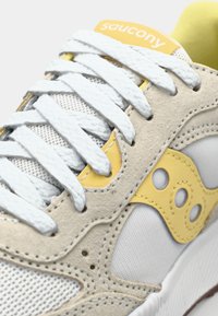 Saucony sneakers with a white mesh and beige suede upper, yellow accents, white laces, and a textured sole design.