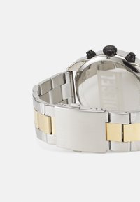 Montre en acier inoxydable argent et or avec une finition polie, un cadran rond et deux boutons poussoirs noirs sur le côté. Comprend un fermoir sécurisé.