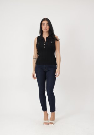 Kobieta stojąca, ubrana w czarny, bezrękawnik z guzikami, ciemne dopasowane dżinsy, białe sandały na obcasie z odkrytymi palcami oraz z widocznym tatuażem na lewym przedramieniu.