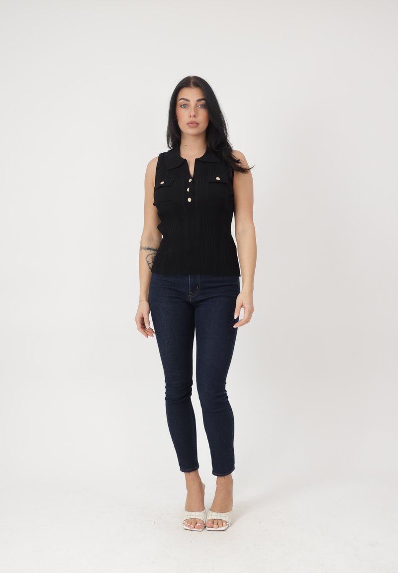 Frau steht, trägt einen ärmellosen schwarzen, geknöpften Oberteil, dunkle enge Jeans, weiße offene High Heels und eine sichtbare Tätowierung am linken Unterarm.