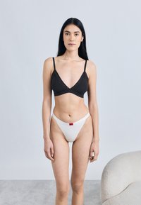 Czarny biustonosz typu bralette z cienkimi ramiączkami, dekolt w kształcie litery V, w połączeniu z białym stringiem z czerwonym logo. Gładka tkanina, minimalistyczny styl.