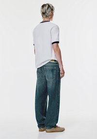 Weißes kurzärmliges Hemd mit marineblauen Akzenten, kombiniert mit lockeren blauen Jeans und beigen Schuhen. Verfügt über einen entspannten Schnitt und ein auffälliges Design der Gesäßtasche.
