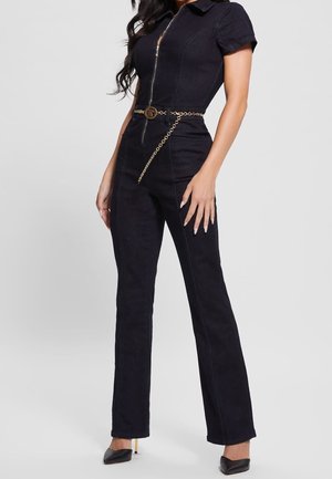 Femme portant une combinaison noire cintrée avec une fermeture éclair à l'avant, des manches courtes et une ceinture chaîne dorée, assortie à des escarpins noirs à bouts pointus.