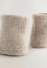 Beige strukturierte Wollslipper mit einem weichen, flauschigen Aussehen, die eine abgerundete Form und einen leicht ausgestellten oberen Rand aufweisen.