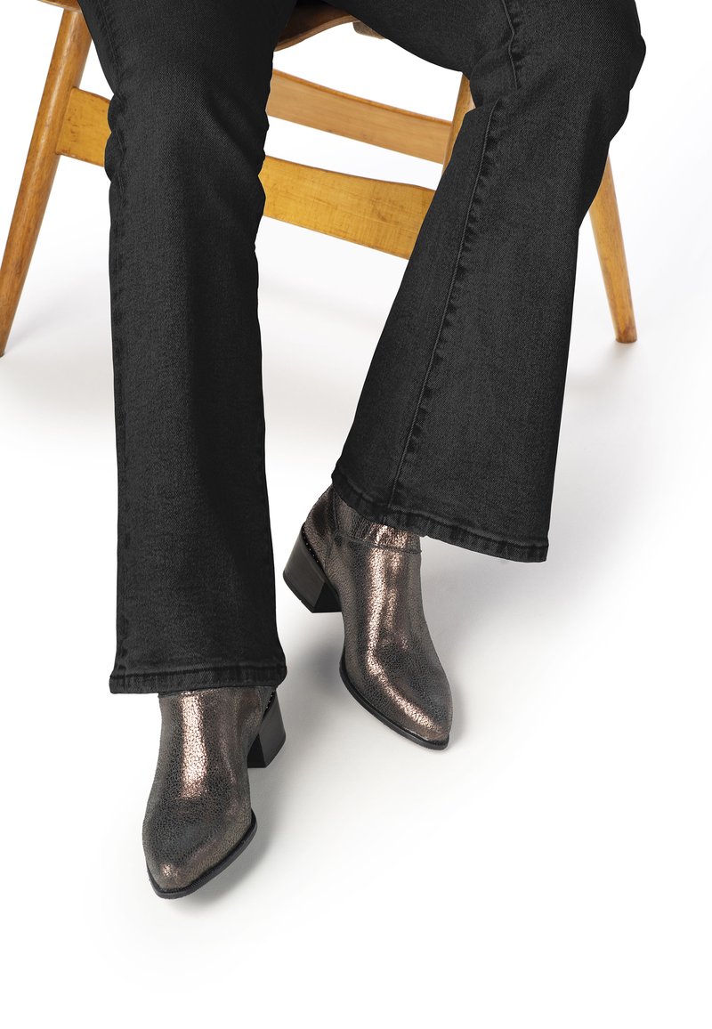 Botas de tobillo de bronce metálico con punta afilada y pequeño tacón cuadrado, combinadas con pantalones de pierna ancha negros. Las botas tienen una superficie texturizada.