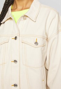 Beige denimjacka med silverbucklor och orange sömnad. Har två bröstfickor och en avslappnad passform. Texturerad tyg.