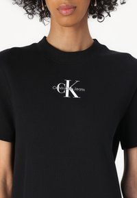 Juodas įvairaus rašto marškinėlis su apvaliu kaklu. Yra baltas Calvin Klein Jeans logotipas ant krūtinės. Minkšta medžiaga ir prigludusi forma.