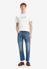 Balta medvilninė marškinė su mėlynu "LEVI'S" logotipu, derinami su šviesiai mėlynomis tiesiomis kelnėmis ir rudos spalvos batais su tekstūruotu paviršiumi.