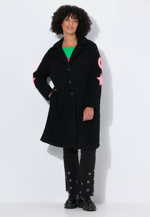 Cappotto di pile nero con colletto, due grandi tasche frontali e chiusura a bottone. Design di stelle rosa sulle maniche. Abbinato a pantaloni neri con grommet.