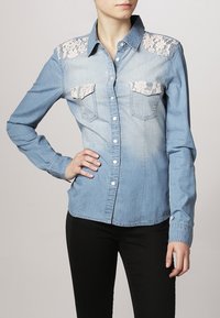 Chemise en denim bleu clair avec manches longues, accents en dentelle aux épaules et deux poches poitrine. Présente un devant à boutons et un ourlet arrondi.