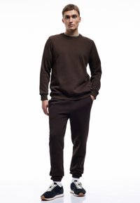 Brun sweatshirt og matchende joggers lavet af blødt stof. Sættet har ribbet manschetter og en rund halsudskæring, parret med mørke sneakers.