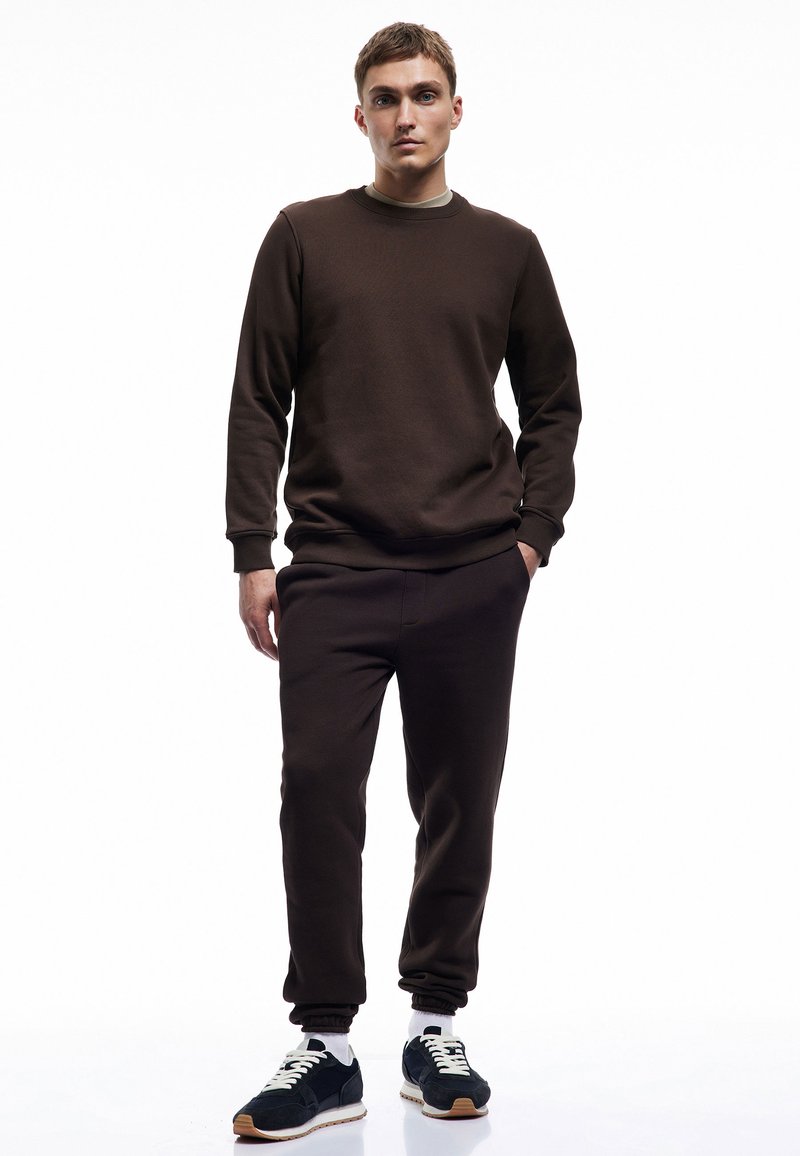 Brun sweatshirt og matchende joggers lavet af blødt stof. Sættet har ribbet manschetter og en rund halsudskæring, parret med mørke sneakers.