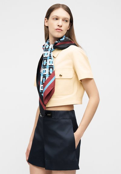 Γυναίκα που φοράει μπεζ κοντό μανίκι σακάκι, μπλε και βουργουνδίας μοτίβο κασκόλ, και σκούρο μπλε σορτς ψηλής μέσης με τα χέρια στις τσέπες.