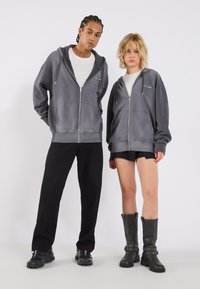Sivi zip-up dukserica s logotipom, nošena s crnim hlačama i čizmama. Model pozira s kratkom i dugom kosom. Jednostavan dizajn, ležeran kroj.
