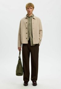 Veste en laine beige, chemise verte à boutons, pantalon large marron et chaussures bordeaux. Sac vert avec détail à cordon en main.