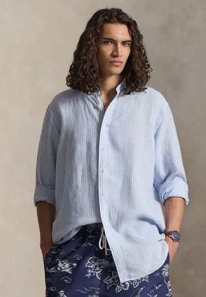 Mann med langt krøllete hår som har på seg en lyseblå stripete skjorte med knapper og marineblå badeshorts med hvite tropiske mønstre.