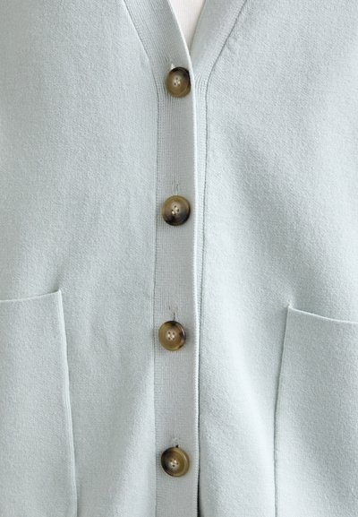 Lys grønn cardigan med v-hals, fem brune knapper og to frontlommer. Laget av myk, strukturert stoff.