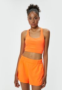 Kirkas oranssi urheilu crop top leveillä olkahihnoilla ja istuvalla muotoilulla, yhdistetty löysiin, joustovyötäröisiin shortseihin samassa värissä.