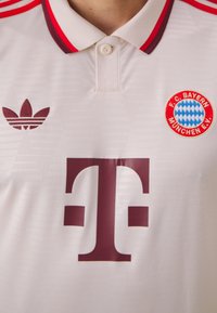 Valkoinen FC Bayern München -jalkapallopaita, jossa on punainen Adidas-logo, seuran vaakuna ja suuri viininpunainen T-Mobilen sponsorilogo rinnassa.