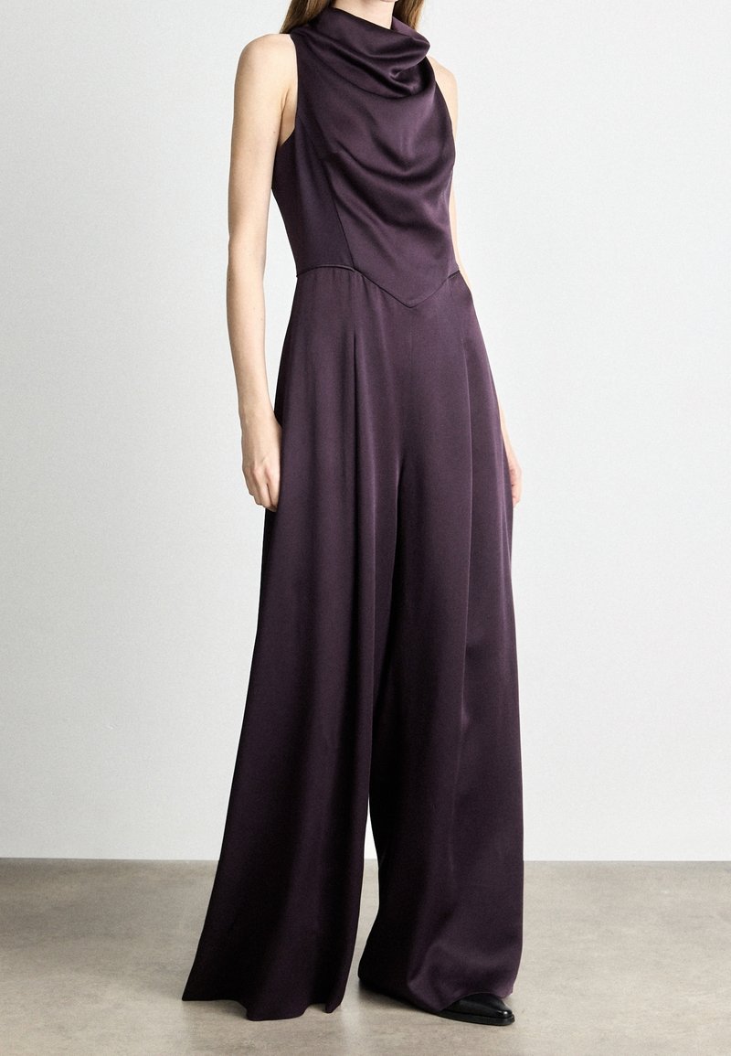 Femme portant une combinaison sans manches violet foncé à jambes larges avec un col drapé, debout sur un sol gris lisse contre un mur blanc.
